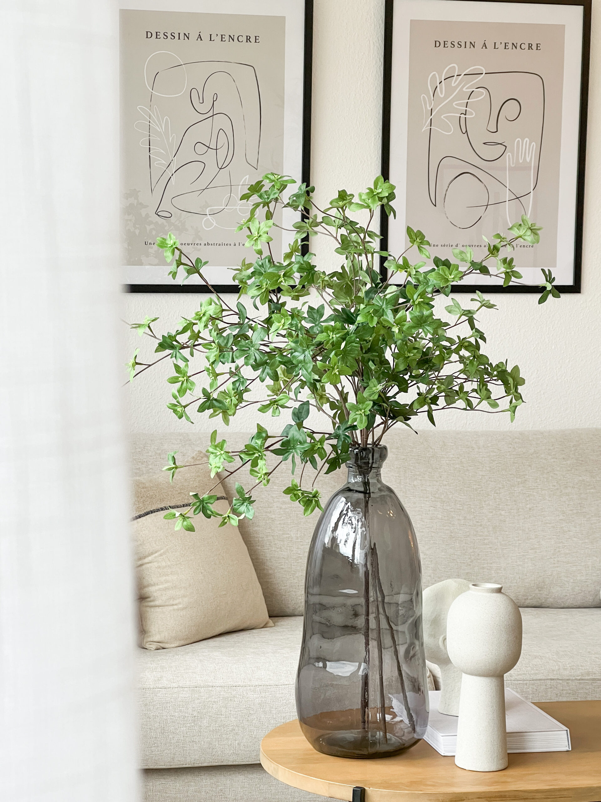 Auf dem Bild ist eine Vase mit Zweigen zu sehen, die auf einem Couchtisch steht. Im Hintergrund sieht man das SOfa und die Wandbilder
